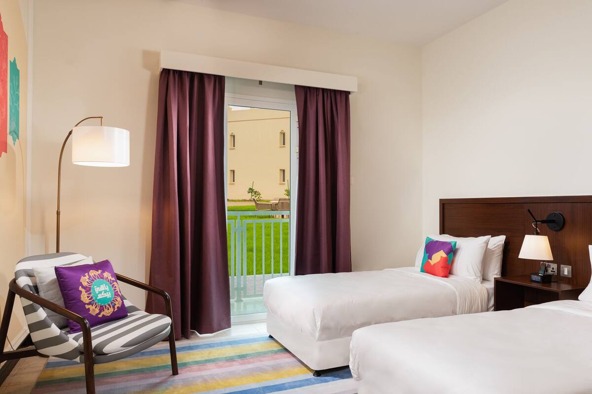Quarto de hotel com duas camas, uma cadeira, uma luminária e vista para uma varanda.