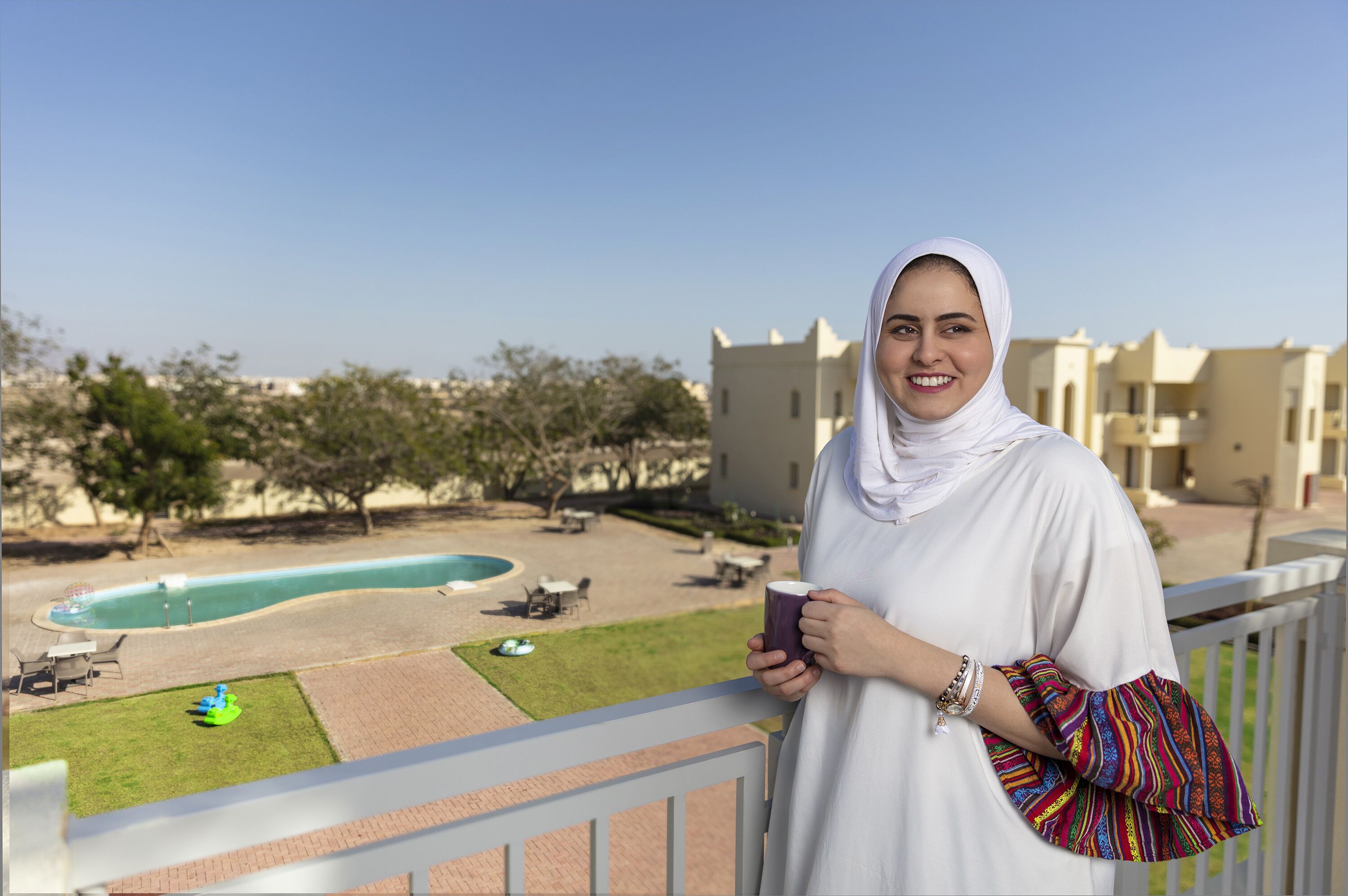 A person standing on a balcony overlooking a swimming pool, with a view of a residential area and buildings in the background.