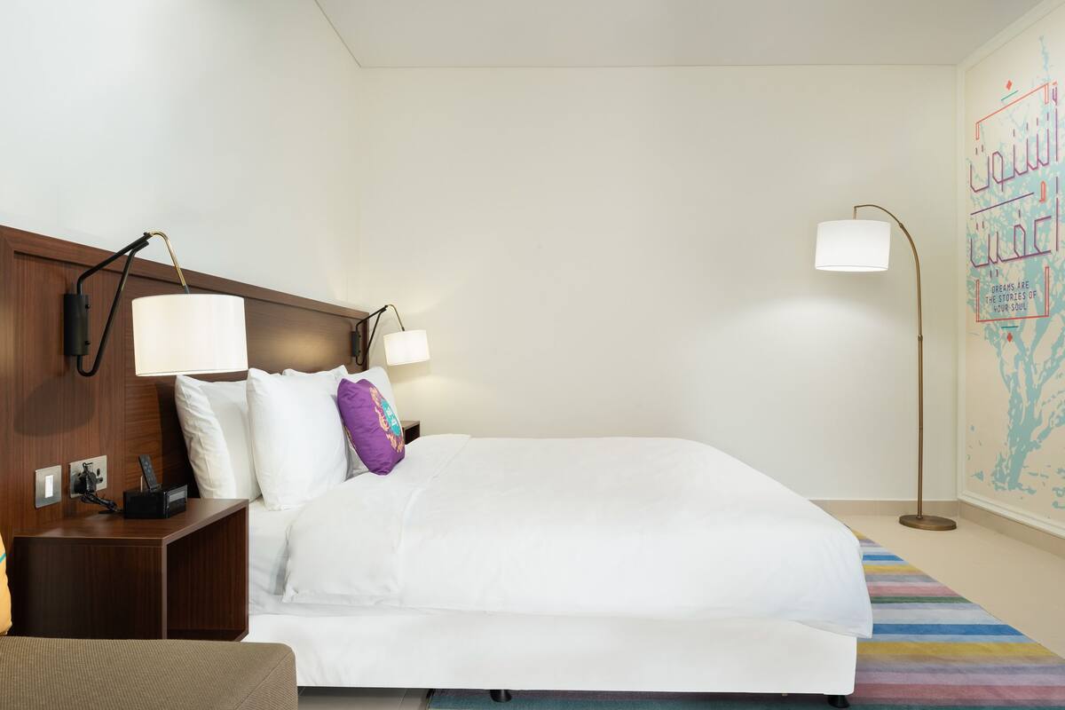Quarto de hotel com cama, mesas de cabeceira, luminárias, tapete listrado e parede com um mural decorativo.