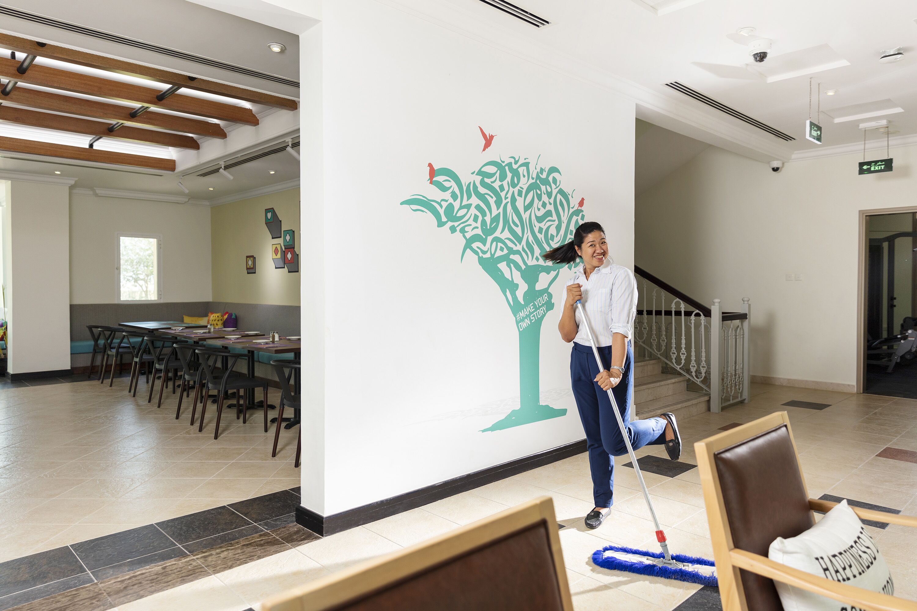 Una persona limpiando el suelo de un pasillo, con un mural decorativo en la pared, una escalera y un comedor al fondo.
