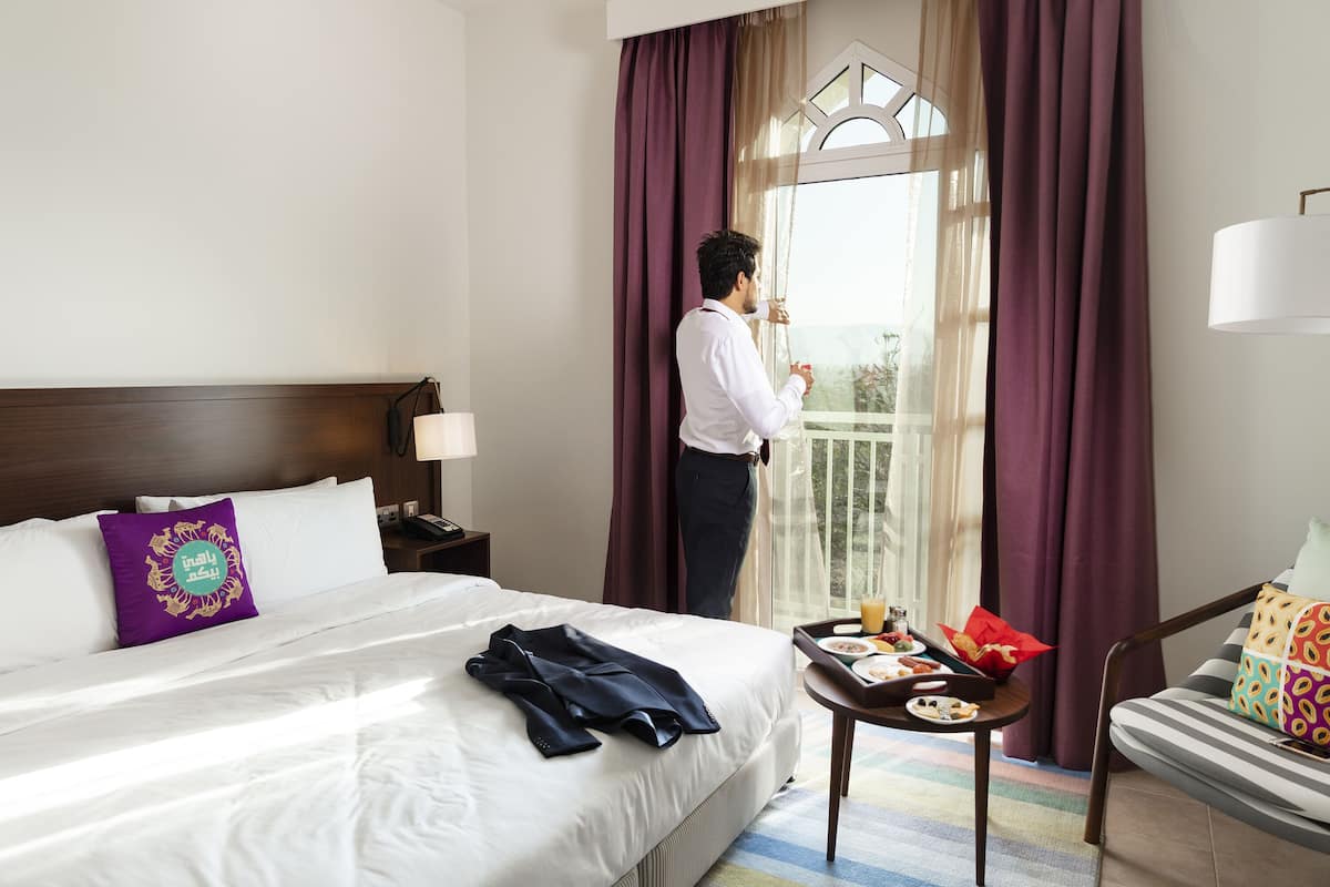Quarto de hotel com cama, mesa de cabeceira, uma mesa com itens de café da manhã e vista para o exterior através de uma janela com cortinas.