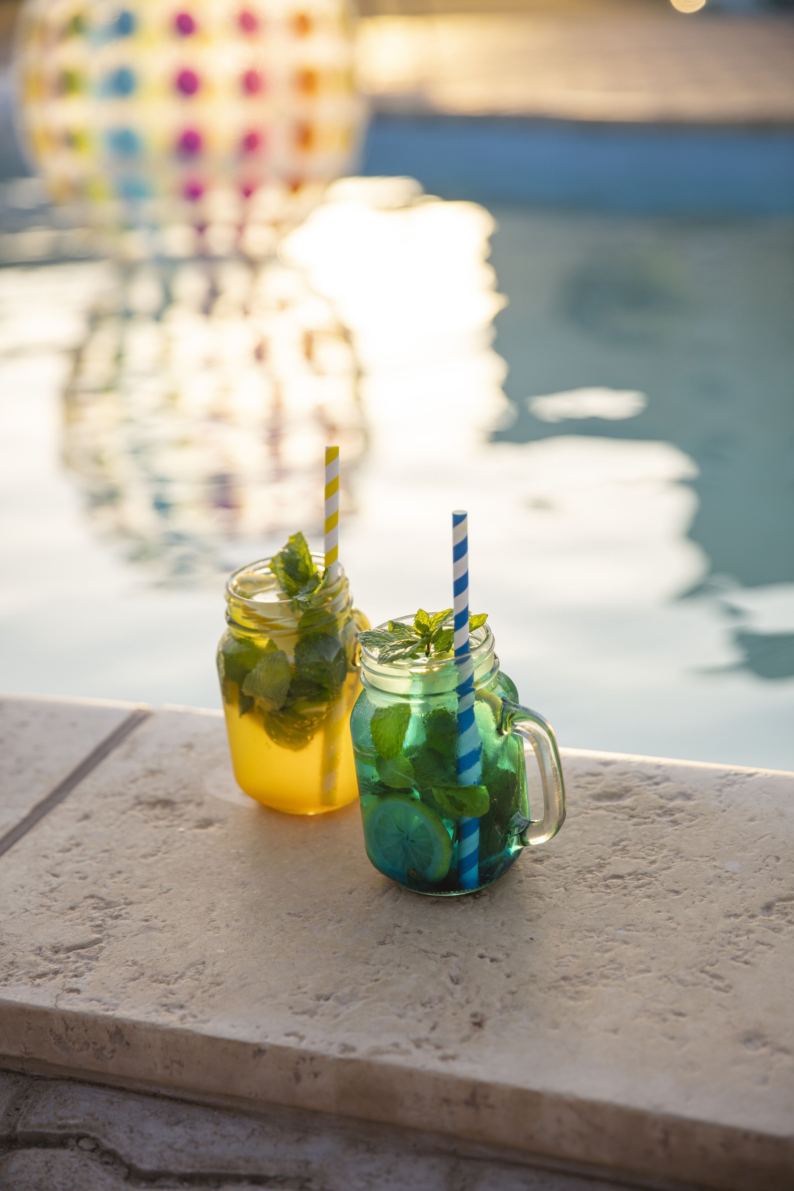 Two mason jars with straws filled with a yellow beverage and mint leaves, placed on a stone ledge by a pool.