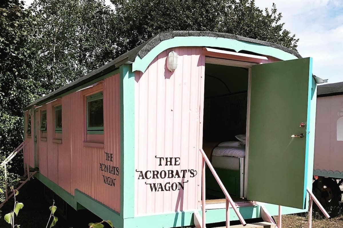A small, pink and green wooden structure labeled 'The Acrobat's Wagon' with a staircase leading up to a bed inside.