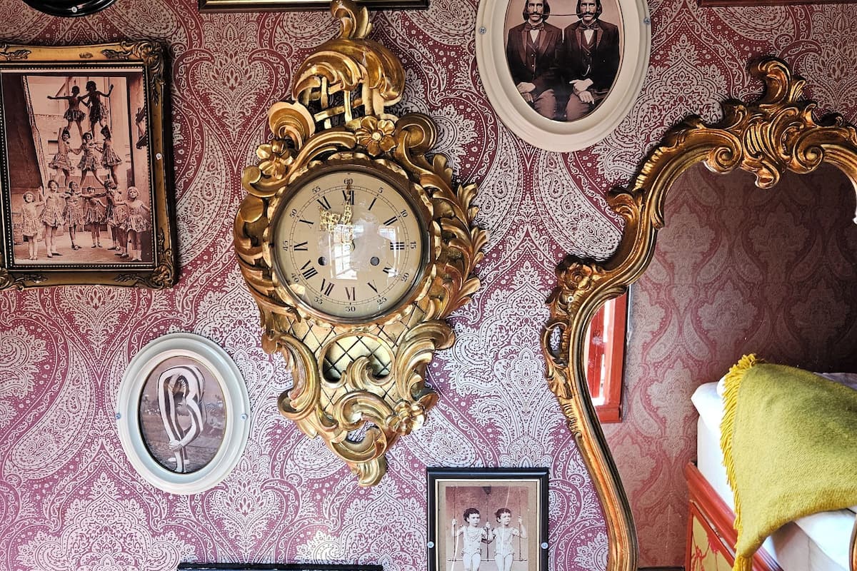 A room with a patterned wallpaper, a large ornate clock, and multiple framed photographs on the wall.
