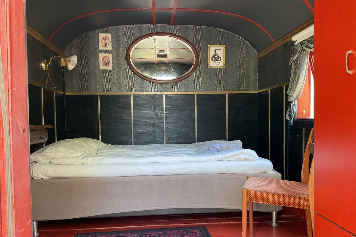A bedroom with a bed, a chair, a red wardrobe, a mirror, and a small rug.