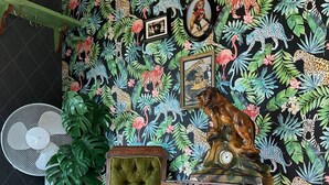 Ein Zimmer mit tropischem Wandpapier, ein Holzstuhl mit grüner Sitzkissen, ein kleiner Tisch mit pinkfarbener Sitzkissen und ein an der Wand befestigter Ventilator.