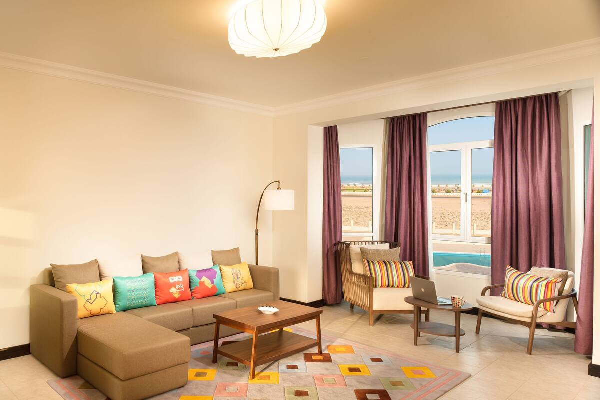 A modern living room with a sofa, armchair, coffee table, and a view of the beach through large windows.