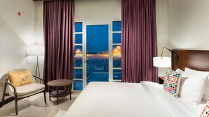 A hotel room with a bed, a chair, a small table, and a view of a beach at night. - Atana Stay Al Ashkarah (Al Ashkharah)