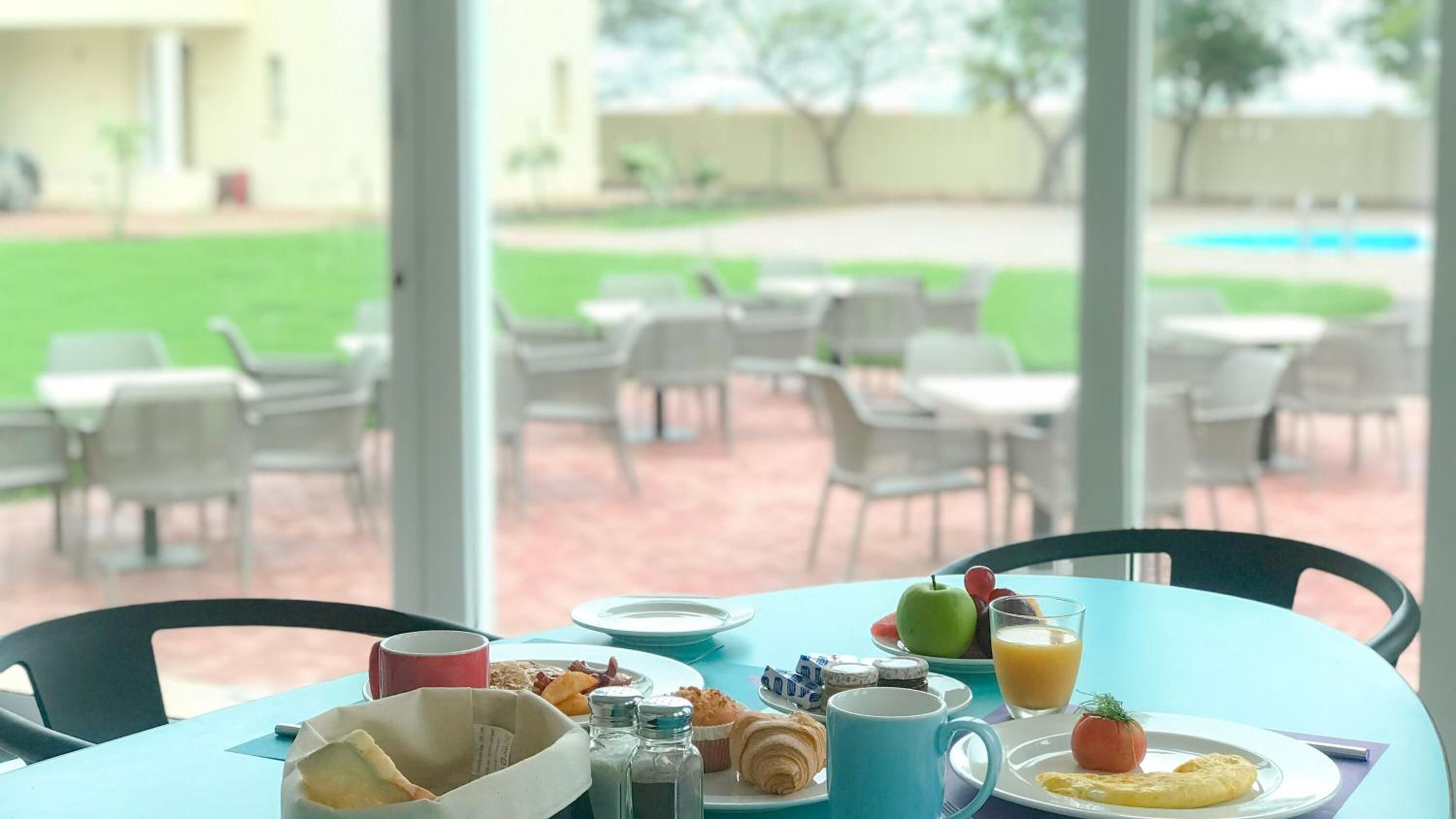 A breakfast table with a variety of food items, including eggs, fruits, and a glass of orange juice, set against a backdrop of outdoor seating and a view of a swimming pool.