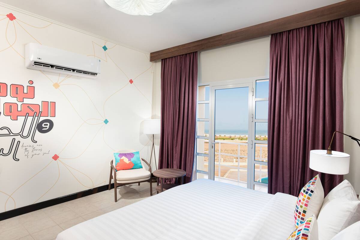 A hotel room with a bed, a chair, a small table, an air conditioning unit, and a view of the beach through a large window.