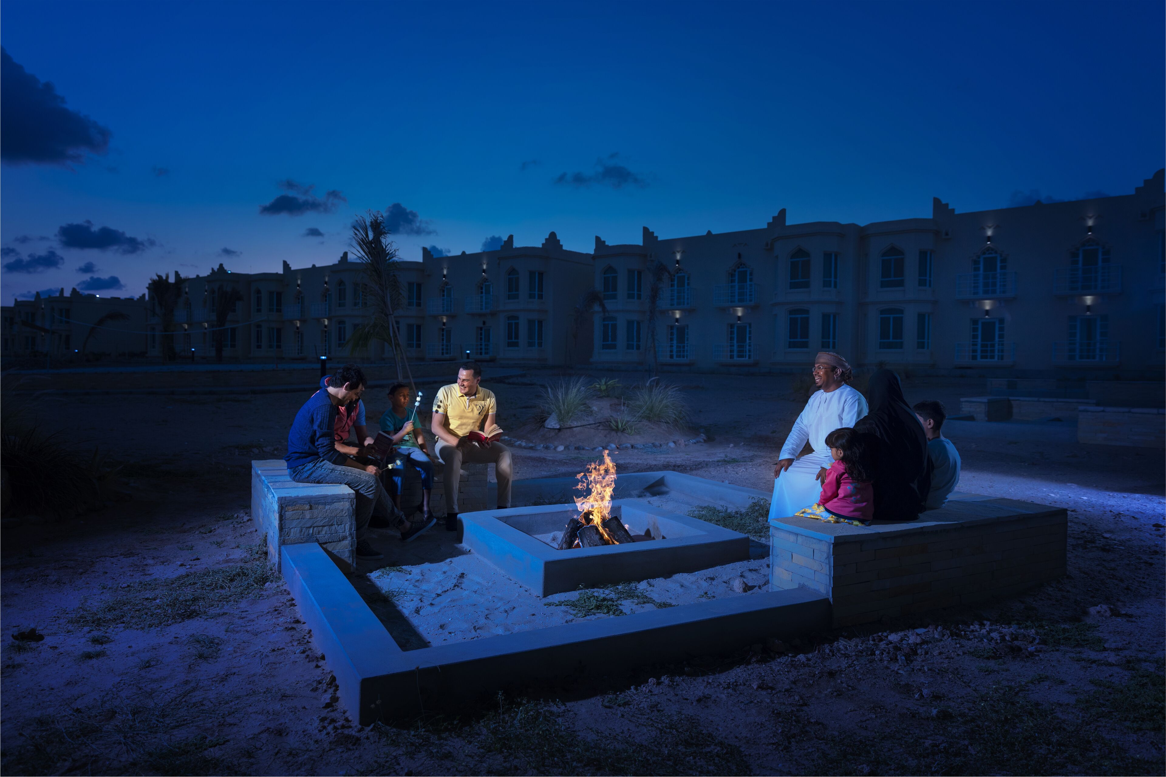 A group of people sitting around a fire pit in an outdoor setting with a large building in the background.