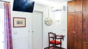 Una habitación con una puerta blanca, un armario de madera, una silla con cojín rojo, una mesita pequeña, un televisor instalado en la pared y un radiador.