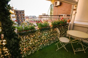 A balcony with a green artificial turf, a white metal table and chair, and potted plants with red flowers. - 96 Guest House (Reggio Calabria)