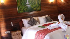 A hotel room with a wooden wall, a painting of a waterfall, a bed with white linens, pillows, and a red blanket. - Putra Bali Villa (Penida Island)