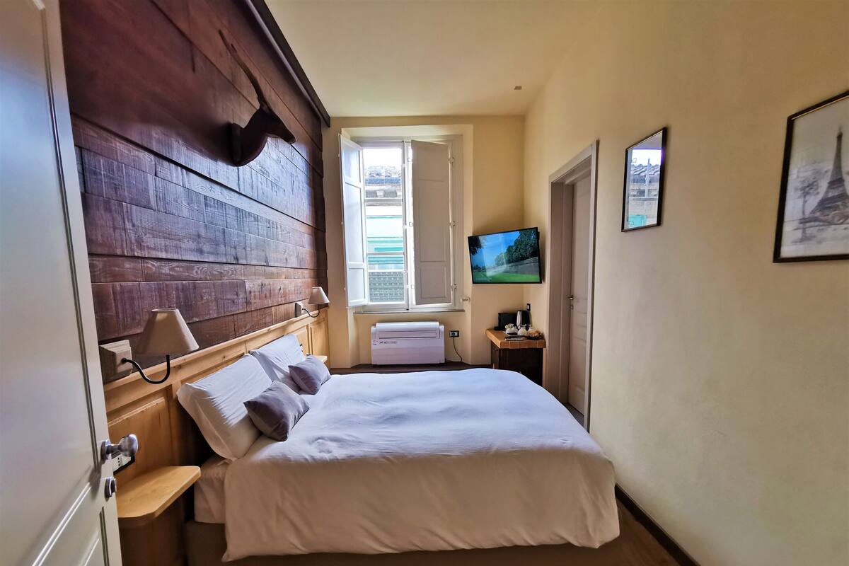 A bedroom with a wooden headboard, a bed with white linens, a TV on the wall, and a window with a view of greenery.