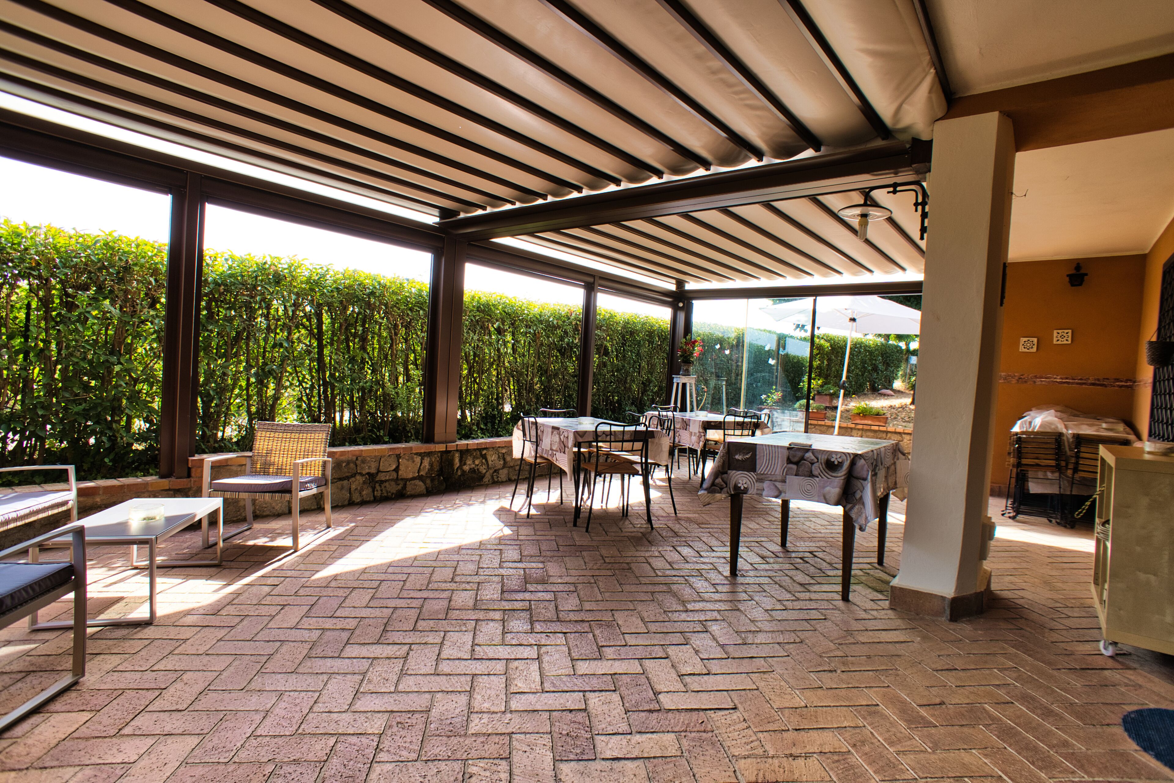 An outdoor patio area with brick flooring, tables, chairs, and a pergola providing shade.