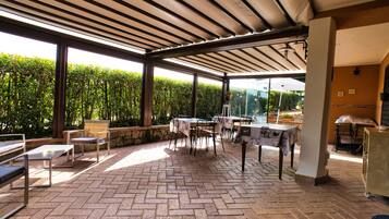 An outdoor patio area with brick flooring, tables, chairs, and a pergola providing shade.