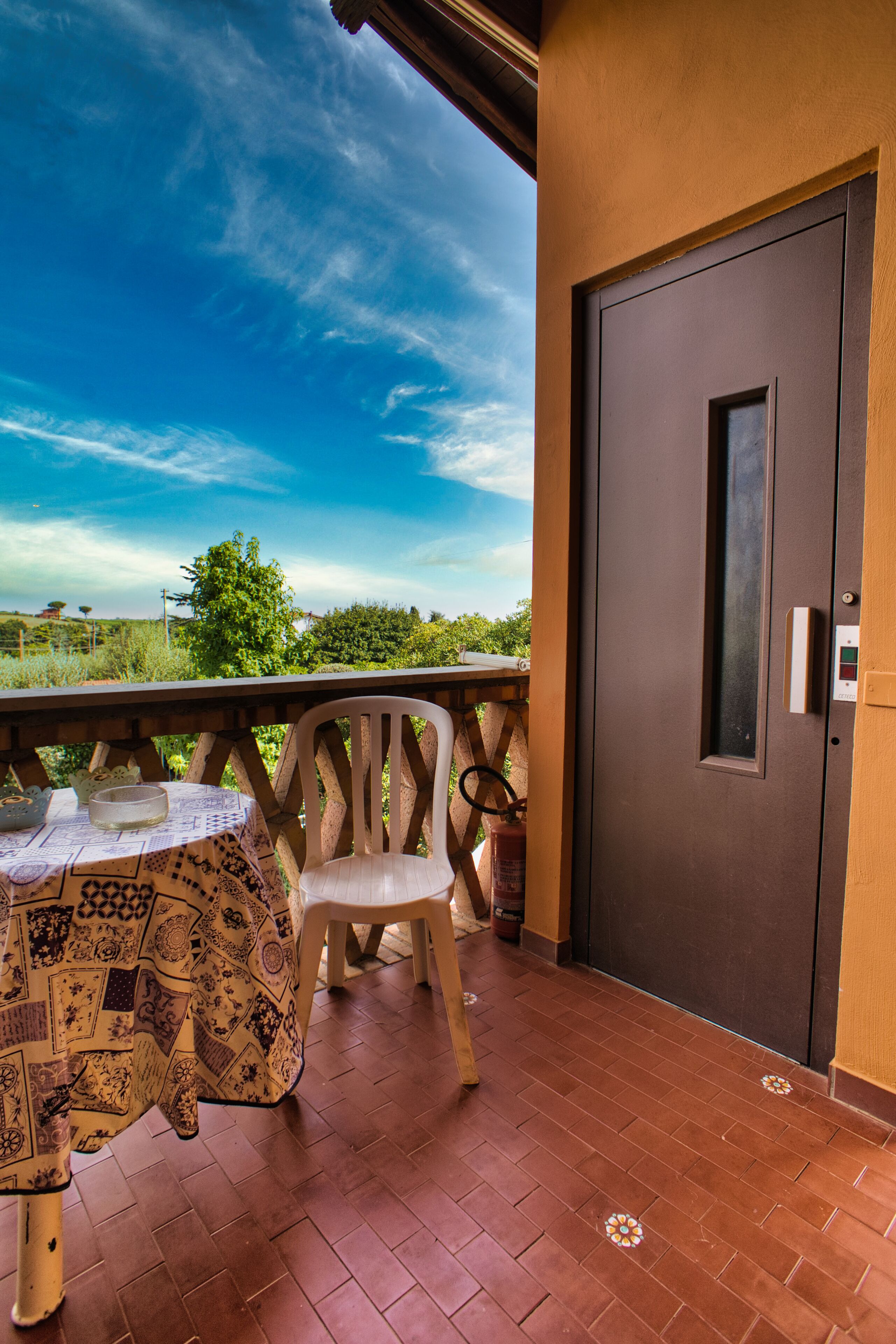 A balcony with a table and chair, a door, and a view of greenery and sky.