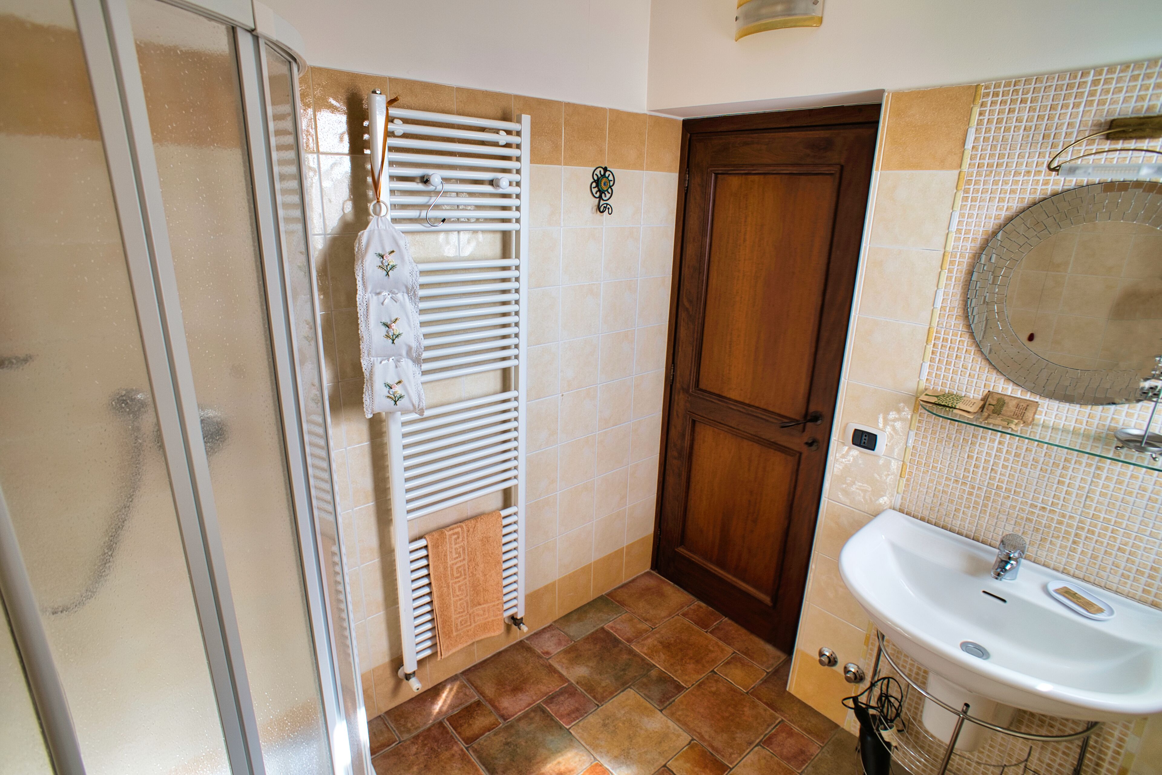 Une salle de bain avec une douche à parois de verre, une porte en bois, un lavabo blanc avec un miroir et un sol carrelé.