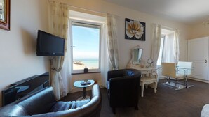 A hotel room with a TV, a sofa, a desk with a mirror, and a view of the beach. - The Golden Hind (St Ives)