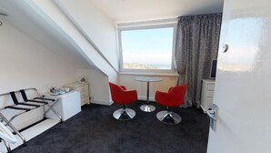 A compact hotel room with a bed, a small desk with a chair, two red chairs around a glass table, a window with a view of the city, and a curtain. - The Golden Hind (St Ives)