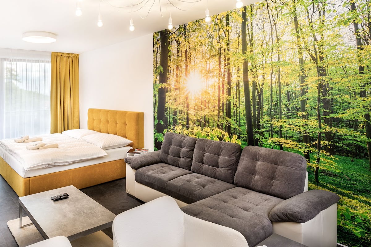 A modern hotel room with a large bed, a grey sofa, and a forest-themed wall mural.
