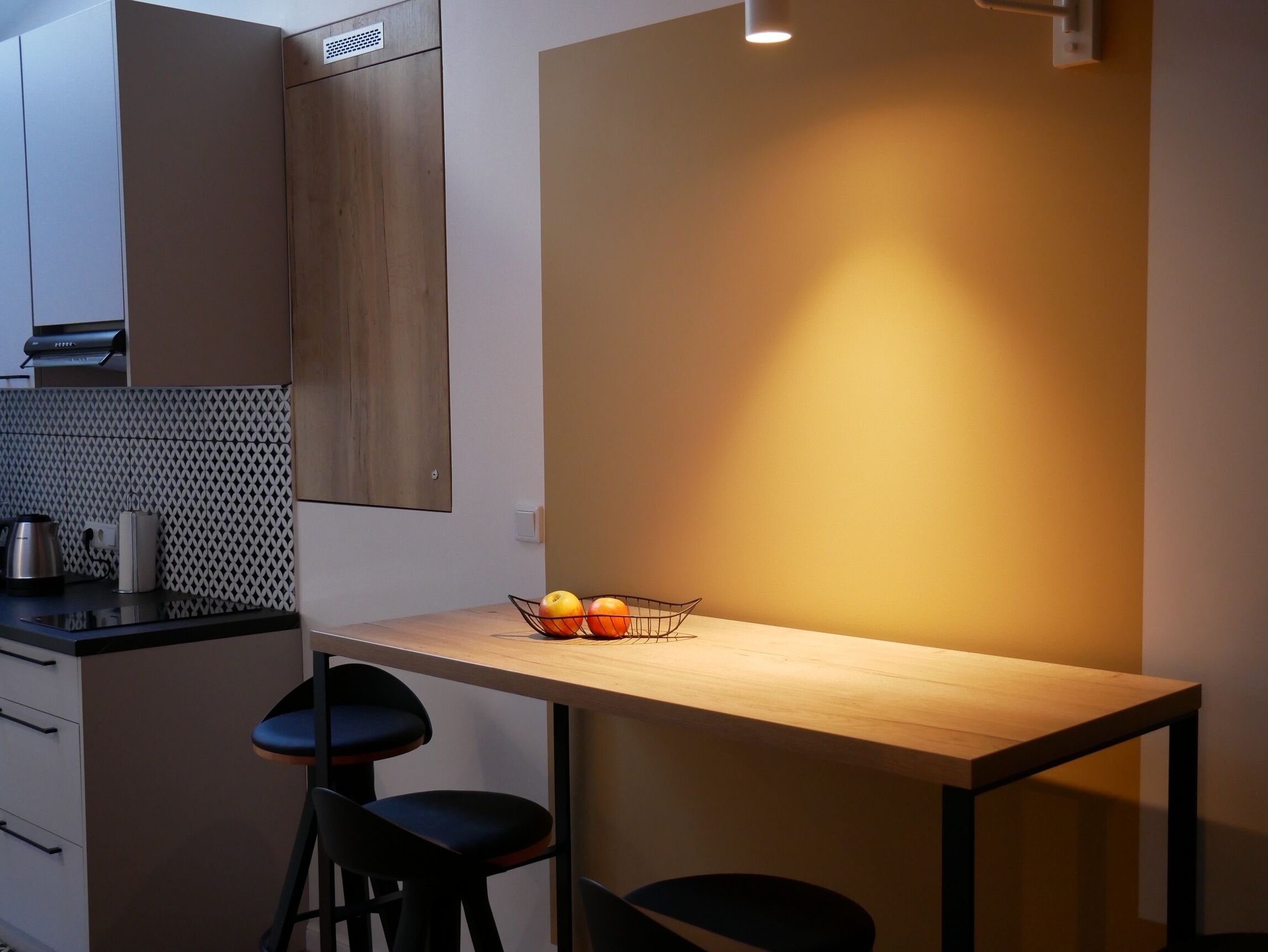 A modern kitchen with a wooden table, two black bar stools, and a fruit bowl containing apples and oranges.