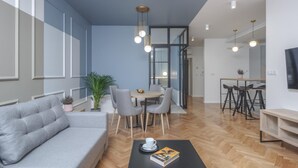 A modern living room with a grey sofa, a coffee table with a book, a dining area with a table and chairs, and a kitchen area with stools and a TV. - Ermine Suites (Kraków)