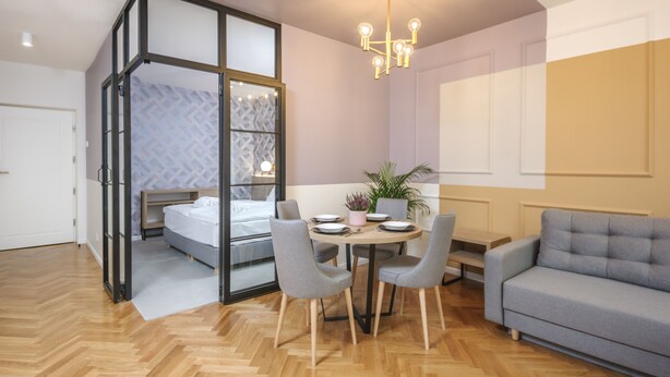 A modern bedroom with a glass partition, a bed, a dining table with chairs, a sofa, and a chandelier. - Ermine Suites (Kraków)