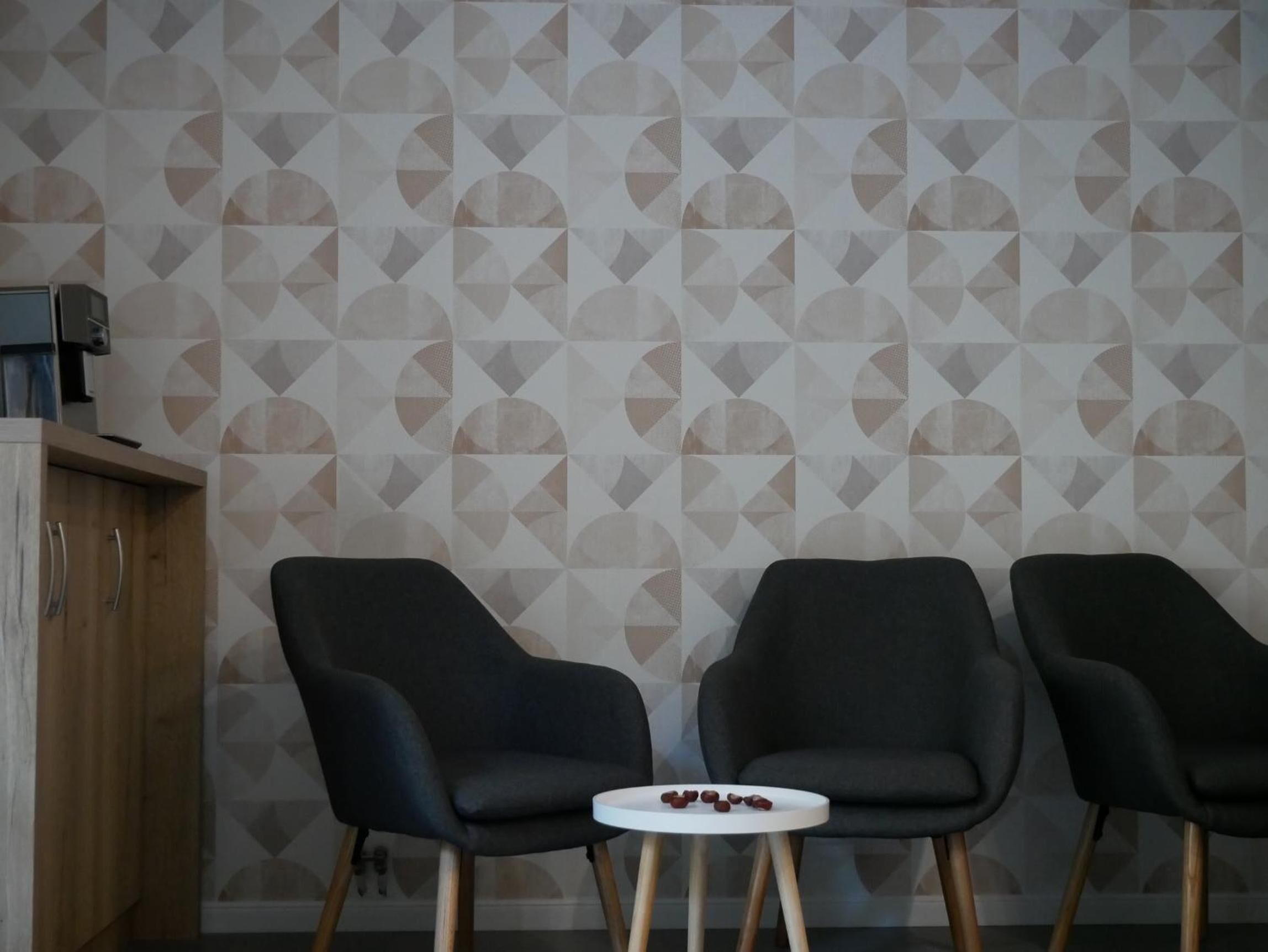 Quarto com papel de parede estampado em padrão geom étrico, duas cadeiras pretas, uma pequena mesa branca com alguns objetos e um armário de madeira.