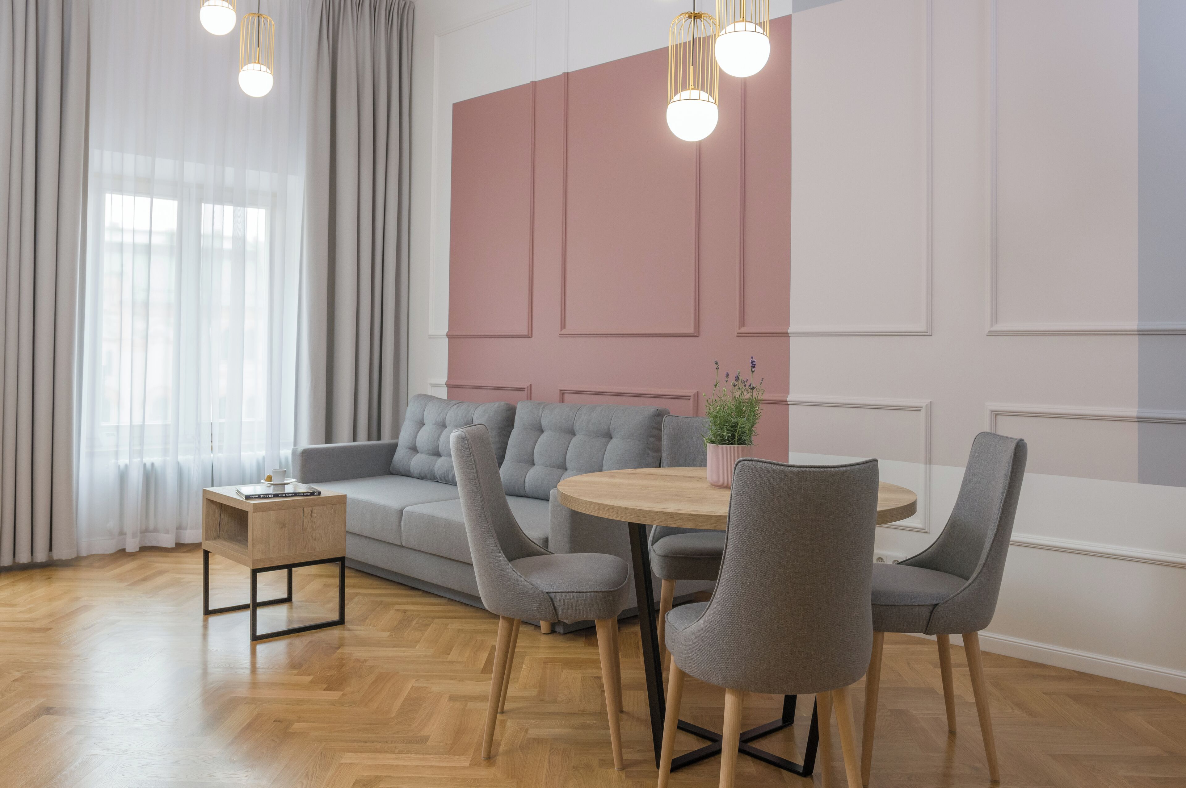 Una sala de estar moderna con un sofá gris, una mesa de centro redonda de madera y sillas grises. Hay una pequeña mesita auxiliar con un libro y una planta. El piso es de madera y hay un ventanal con cortinas de gasa.