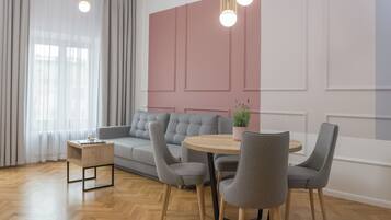 Una sala de estar moderna con un sofá gris, una mesa de centro redonda de madera y sillas grises. Hay una pequeña mesita auxiliar con un libro y una planta. El piso es de madera y hay un ventanal con cortinas de gasa.