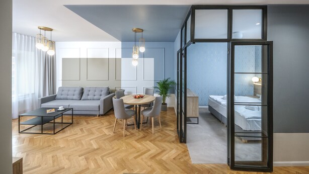 A modern living room with a sofa, dining table, and chairs, a separate bedroom with a bed, and a large window with curtains. - Ermine Suites (Kraków)