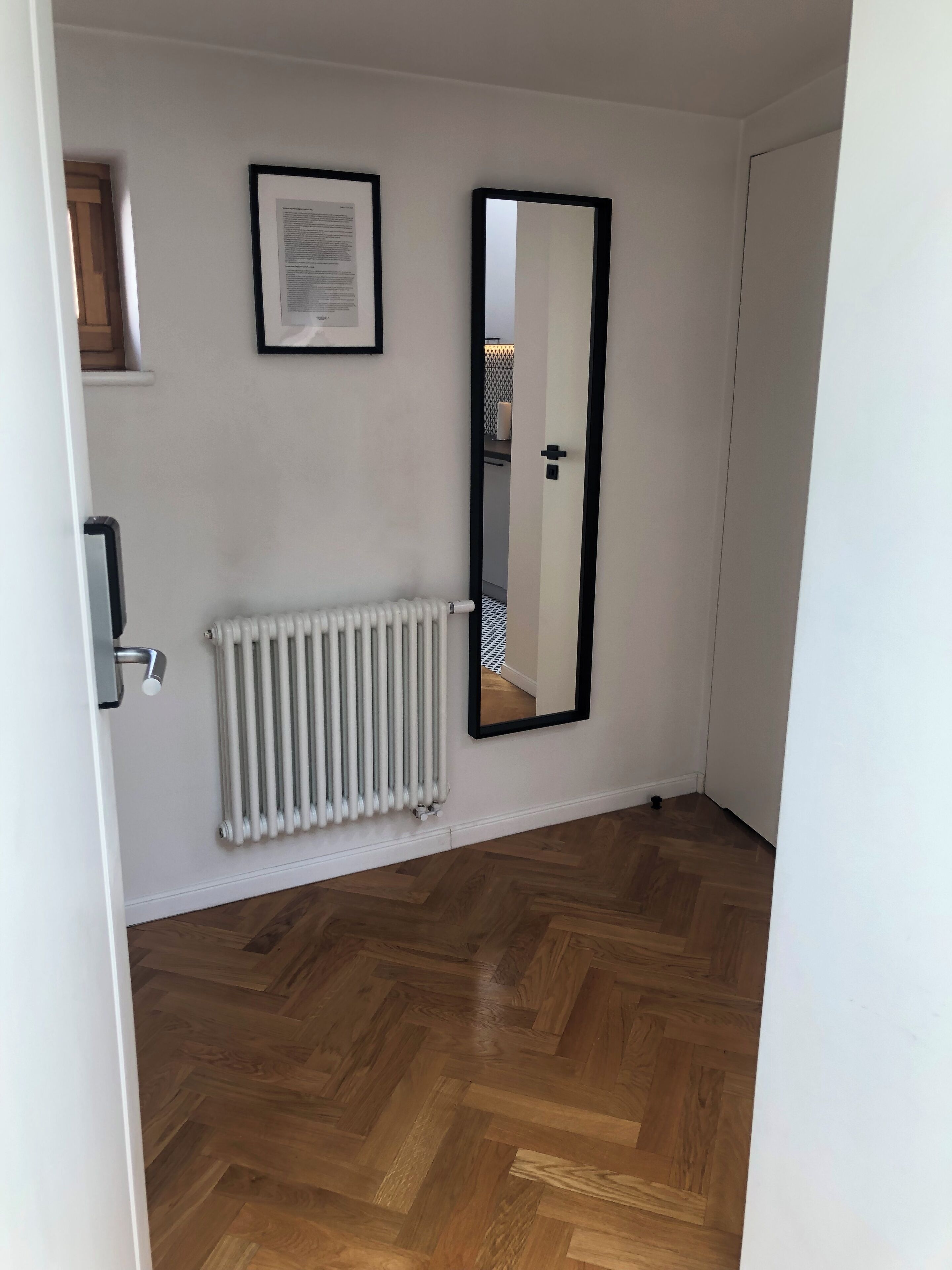 Ein Flur mit Heizkörper, einem gerahmten Dokument an der Wand und einem Spiegel, der eine Tür reflektiert.
