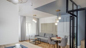 A modern living room with a sofa, coffee table, dining table, and chairs. There is a large window with curtains, a ceiling light fixture, and a door with glass panels. - Ermine Suites (Kraków)