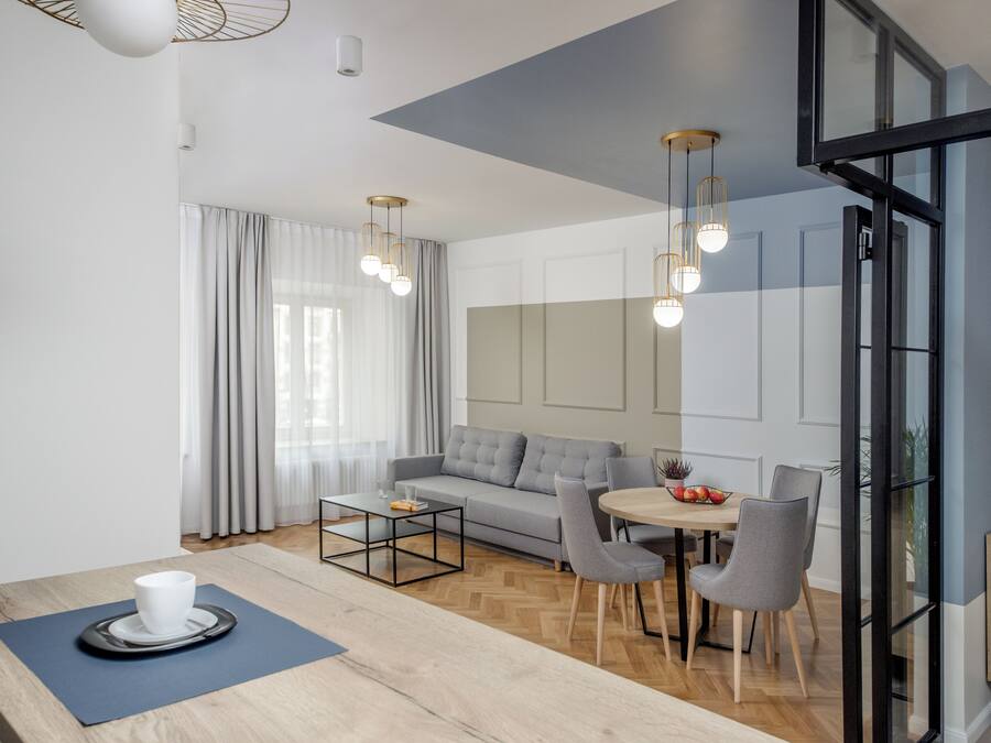 Een moderne woonkamer met een bank, salontafel, eettafel en stoelen. Er is een groot raam met gordijnen, een plafondlamp en een deur met glaspanelen.