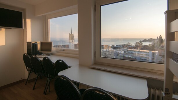 A room with a large window offering a view of a cityscape and a body of water. There is a desk with a computer and chairs. - Skyhostel Friedrichshafen (Friedrichshafen)