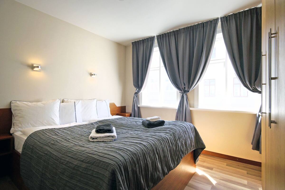 Uma cama bem arrumada com colcha listrada, uma mesa de cabeceira com abajur e uma janela com cortinas.