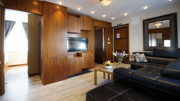 A modern living room with wooden paneling, a flat-screen TV, a black leather sofa, and a small coffee table with a bowl of fruit.
