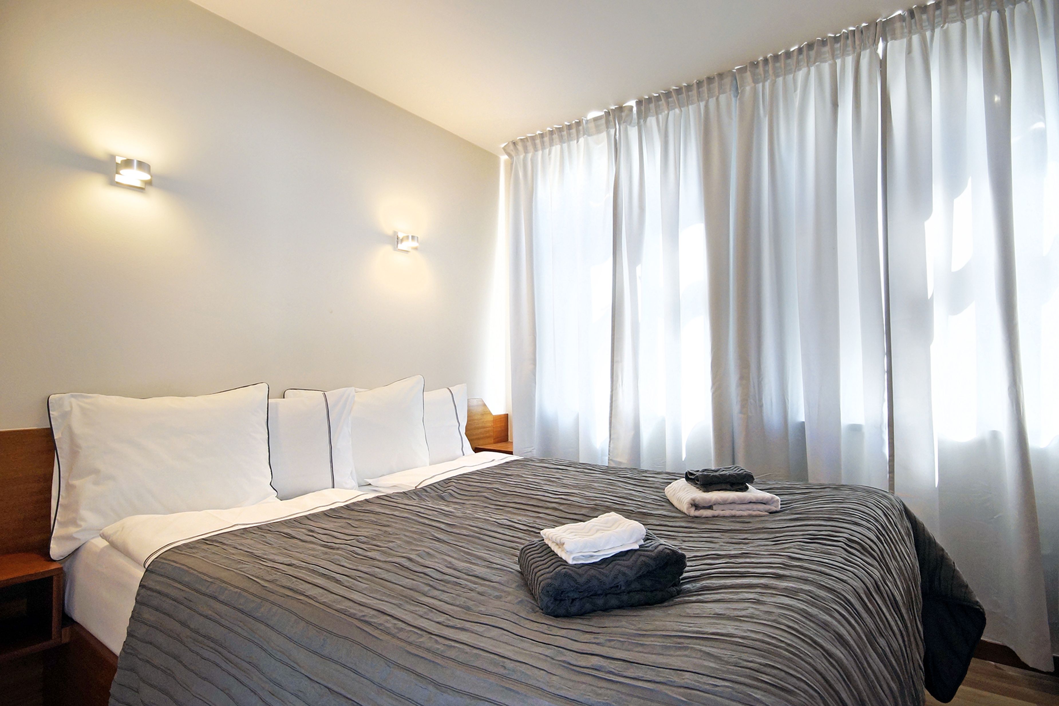 Une chambre d’hôtel avec un grand lit, du linge de lit blanc, une tête de lit en bois et une fenêtre ornée de rideaux légers.