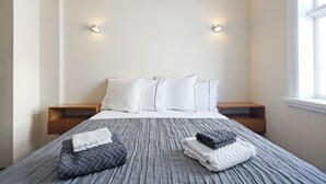 A neatly made bed with pillows and folded blankets, bedside tables, and wall-mounted lights in a simple, clean room.