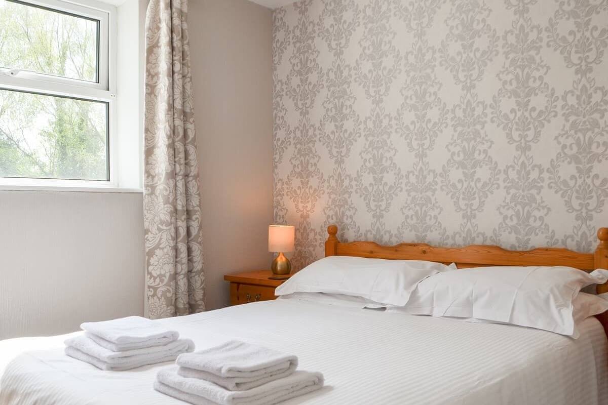 A neatly made bed with white linens and towels, a wooden headboard, a nightstand with a lamp, and a window with curtains.