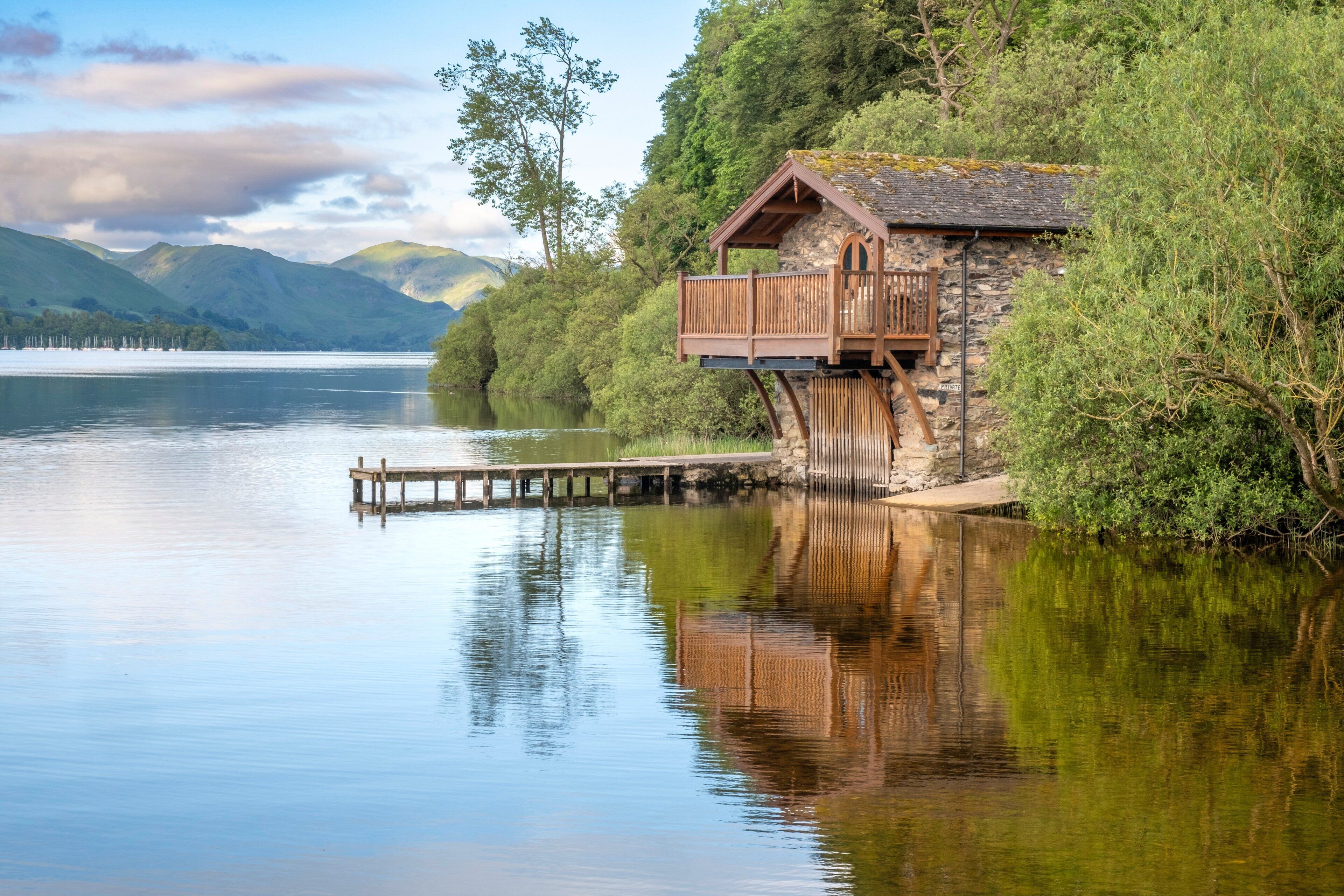 Une cabane en pierre dotée d’un balcon en bois qui surplombe un lac paisible, entourée d’une végétation luxuriante et avec des montagnes en arrière-plan.