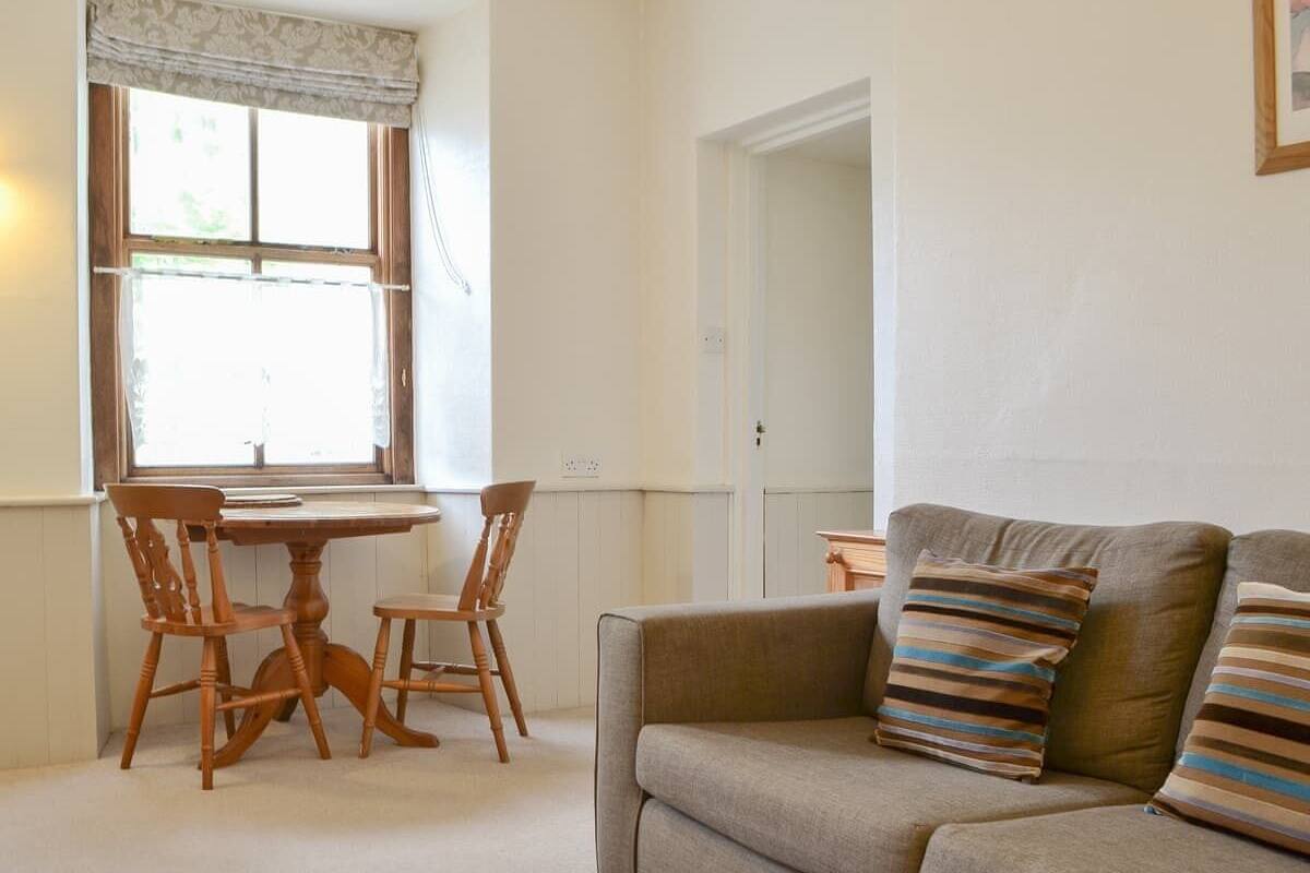 A living room with a beige sofa, striped cushions, a wooden dining table with chairs, and a window with curtains.