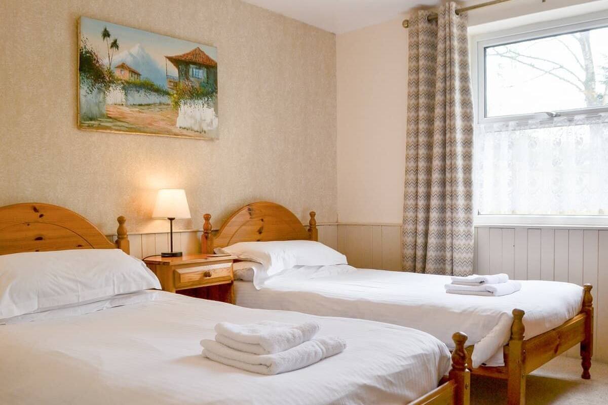 A hotel room with two wooden beds, a nightstand with a lamp, a painting on the wall, and a window with curtains.
