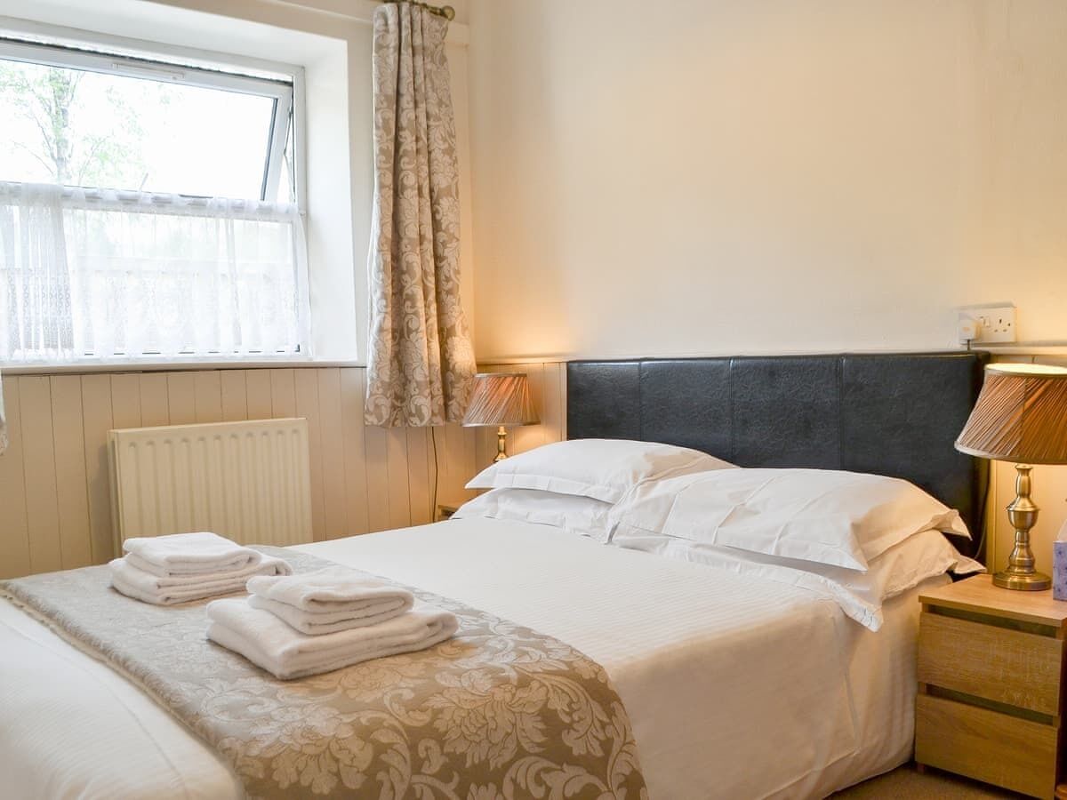 A neatly made bed with white linens and a patterned throw, a bedside lamp, and a window with curtains.