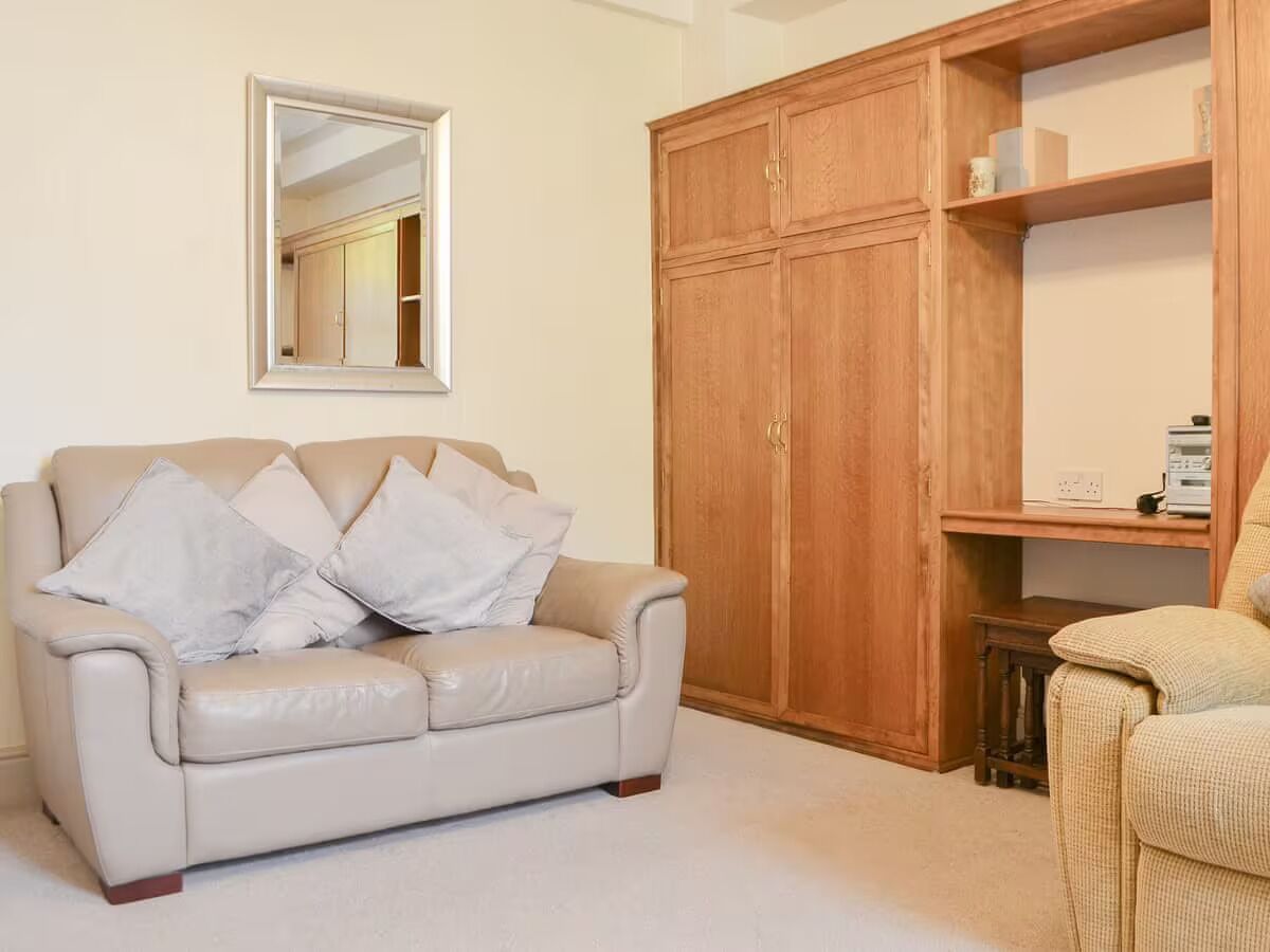 A living room with a beige leather sofa, a wooden wardrobe, and a small table with a lamp.