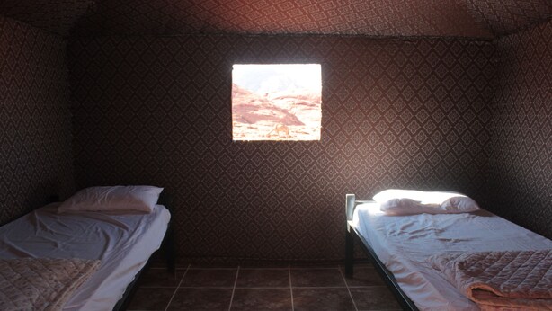 A room with two beds, patterned walls, and a window with a view of a mountainous landscape. - Bedouin Nomads Adventures (Wadi Rum)
