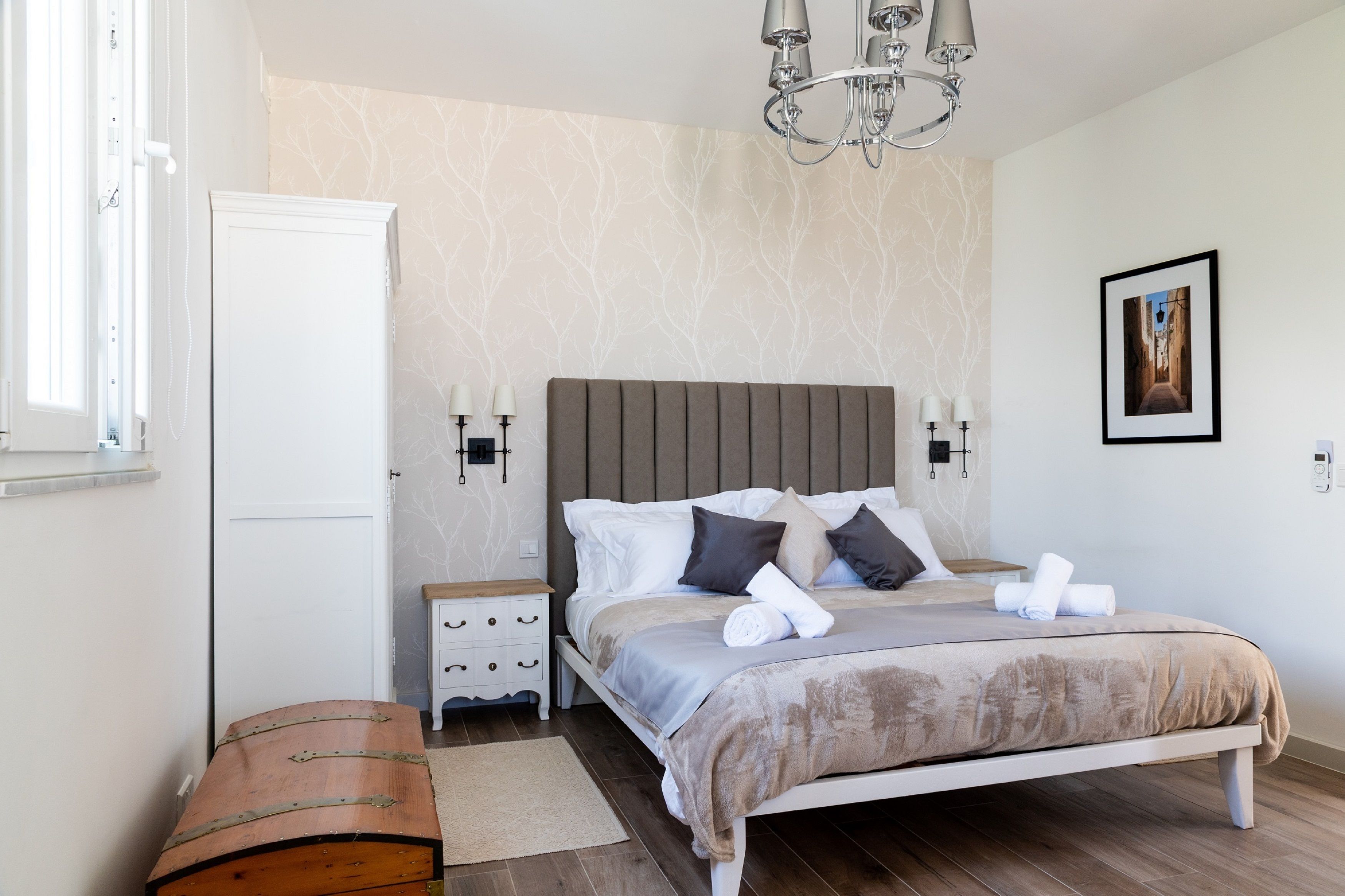 A bedroom with a bed, bedside tables, a wardrobe, a wooden bench, and a chandelier.