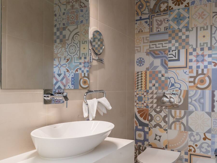 A modern bathroom with a large white sink, a toilet, and a mirror. The walls are decorated with patterned tiles.
